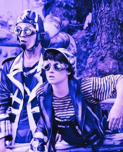 Dos personas con gafas y sombreros vintage mirando hacia adelante, apoyados en una valla de madera, con un filtro azul.